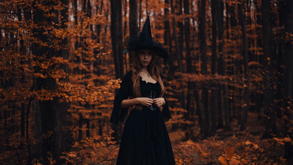 a woman in a black dress and hat standing in a forest