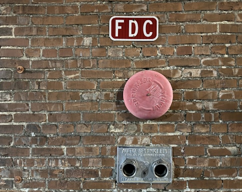 A brick wall features fire department connections including a red 'FDC' sign at the top, a circular pink 'Sprinkler Alarm' cover in the middle, and a metal auto sprinkler connection with two openings at the bottom.