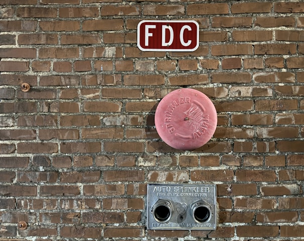 A brick wall features fire department connections including a red 'FDC' sign at the top, a circular pink 'Sprinkler Alarm' cover in the middle, and a metal auto sprinkler connection with two openings at the bottom.