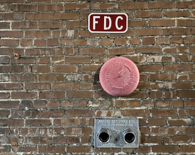 A brick wall features fire department connections including a red 'FDC' sign at the top, a circular pink 'Sprinkler Alarm' cover in the middle, and a metal auto sprinkler connection with two openings at the bottom.