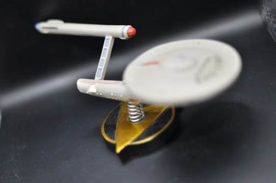 A miniature model of a spaceship is displayed against a dark background. The spaceship has a sleek design with a saucer section and elongated nacelles, positioned on a spiral-shaped stand with a black and gold base.