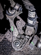 A close-up view of a mechanical part, likely from a vehicle, resting on a rough, asphalt surface. The part appears worn and dirty, with visible scratches and markings. There is a small label with text and a warning attached to one of the components. The part consists of metal and rubber elements, with some bolts and joints visible. Green markings in the shape of the letter 'X' are visible on the part.