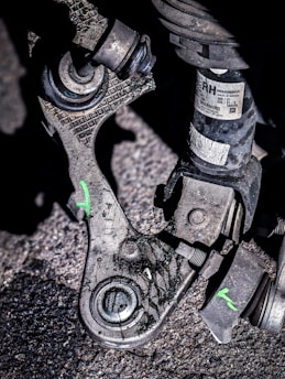 A close-up view of a mechanical part, likely from a vehicle, resting on a rough, asphalt surface. The part appears worn and dirty, with visible scratches and markings. There is a small label with text and a warning attached to one of the components. The part consists of metal and rubber elements, with some bolts and joints visible. Green markings in the shape of the letter 'X' are visible on the part.