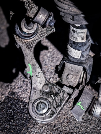 A close-up view of a mechanical part, likely from a vehicle, resting on a rough, asphalt surface. The part appears worn and dirty, with visible scratches and markings. There is a small label with text and a warning attached to one of the components. The part consists of metal and rubber elements, with some bolts and joints visible. Green markings in the shape of the letter 'X' are visible on the part.