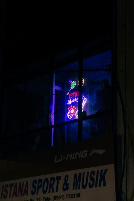 A neon sign in a window displays vibrant colors with tennis sporting motifs and text, which appears above a business sign advertising sporting goods and music. The setting is dimly lit, enhancing the glow of the neon signage.