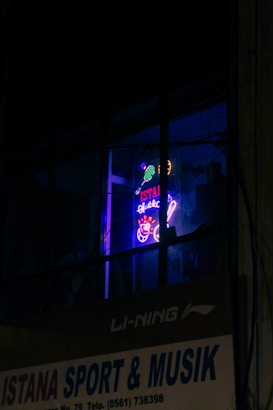 A neon sign in a window displays vibrant colors with tennis sporting motifs and text, which appears above a business sign advertising sporting goods and music. The setting is dimly lit, enhancing the glow of the neon signage.