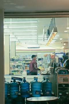 A person is standing at the checkout counter in a well-lit convenience store. Shelves filled with various products are visible in the background. Several large blue water containers are stacked near the entrance.