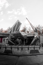 A large sci-fi spaceship with angular and mechanical features is stationed on a concrete platform. The environment includes industrial structures and background foliage of trees, with a light, overcast sky.