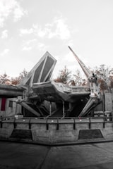 A large sci-fi spaceship with angular and mechanical features is stationed on a concrete platform. The environment includes industrial structures and background foliage of trees, with a light, overcast sky.