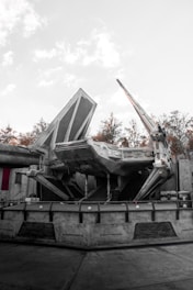 A large sci-fi spaceship with angular and mechanical features is stationed on a concrete platform. The environment includes industrial structures and background foliage of trees, with a light, overcast sky.