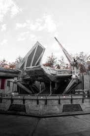 A large sci-fi spaceship with angular and mechanical features is stationed on a concrete platform. The environment includes industrial structures and background foliage of trees, with a light, overcast sky.