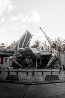 A large sci-fi spaceship with angular and mechanical features is stationed on a concrete platform. The environment includes industrial structures and background foliage of trees, with a light, overcast sky.