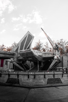 A large sci-fi spaceship with angular and mechanical features is stationed on a concrete platform. The environment includes industrial structures and background foliage of trees, with a light, overcast sky.