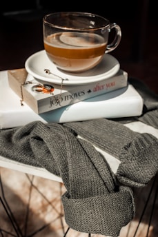 A glass cup of coffee rests on a saucer placed on top of a book. The book, titled 'After You' by Jojo Moyes, has a delicate necklace draped over it. Surrounding the setup is a cozy, knitted gray sweater draped over a round, white table.