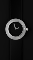 A modern, minimalistic wall clock with a round metallic frame and a black face. The clock has simple, straight hands and no numbers. It is set against a black background, which blends seamlessly with the clock's face.