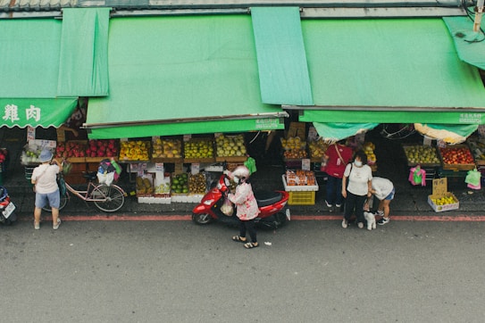 A street market scene with several people shopping for fresh produce. There are green awnings covering the market stalls, which display a variety of fruits. A person is seen walking a dog, while others are browsing or purchasing goods. Bicycles and motor scooters are parked along the street.