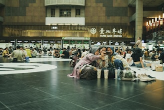 A large indoor space filled with numerous people sitting on the floor, some engaged in conversation while others are looking at their phones. In the foreground, three young women are taking a selfie together, sitting on the black tiled floor. They have various items such as bags and drinks placed nearby. The background features signage indicating 'Taipei Main Station', with digital departure boards and a variety of lights and escalators visible, contributing to a bustling environment.