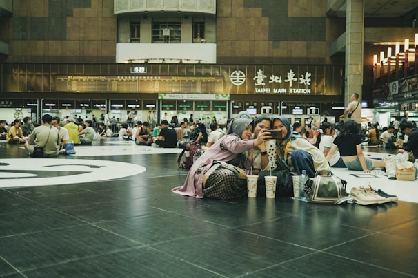 A large indoor space filled with numerous people sitting on the floor, some engaged in conversation while others are looking at their phones. In the foreground, three young women are taking a selfie together, sitting on the black tiled floor. They have various items such as bags and drinks placed nearby. The background features signage indicating 'Taipei Main Station', with digital departure boards and a variety of lights and escalators visible, contributing to a bustling environment.