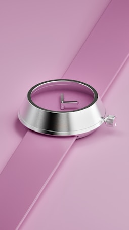 A modern, minimalistic silver clock is placed on a pink background. The clock has a metallic finish and sleek design, with simple hour and minute hands pointing to the time. The overall aesthetic is clean and stylish.