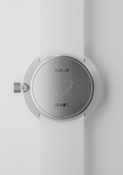 A close-up view of a silver wristwatch with a white strap. The back of the watch shows the outline of Nigeria, with the text 'NAIJA MADE'. The watch's crown is visible on the left side of the image.