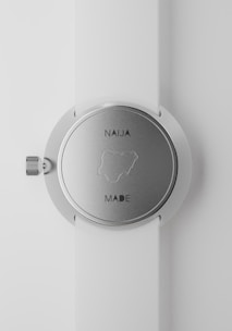 A close-up view of a silver wristwatch with a white strap. The back of the watch shows the outline of Nigeria, with the text 'NAIJA MADE'. The watch's crown is visible on the left side of the image.