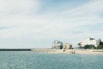A serene beach scene with calm turquoise waters, a sandy shore, and a few people swimming and walking along the beach. Several buildings with distinct architectural styles are visible along the shoreline, surrounded by a few palm trees. The sky above is expansive and mostly clear with a subtle gradient.