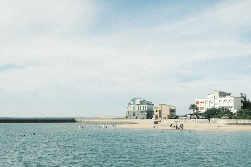 A serene beach scene with calm turquoise waters, a sandy shore, and a few people swimming and walking along the beach. Several buildings with distinct architectural styles are visible along the shoreline, surrounded by a few palm trees. The sky above is expansive and mostly clear with a subtle gradient.