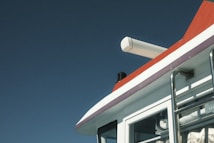 A close-up of a boat's cabin features a clean, modern design with a large pipe or exhaust on the rooftop and stainless steel railing. The cabin is mostly white with a striking red accent at the top, set against a clear blue sky.