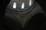 A detailed architectural interior scene with tall arched windows allowing light to enter. The intricate stone carvings include an angelic figure with wings prominently displayed against the curved surfaces of the structure.