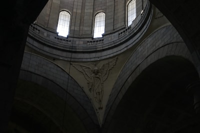 A detailed architectural interior scene with tall arched windows allowing light to enter. The intricate stone carvings include an angelic figure with wings prominently displayed against the curved surfaces of the structure.