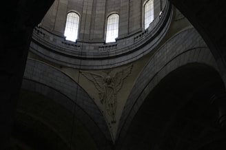 A detailed architectural interior scene with tall arched windows allowing light to enter. The intricate stone carvings include an angelic figure with wings prominently displayed against the curved surfaces of the structure.