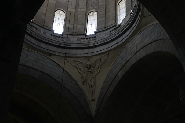 A detailed architectural interior scene with tall arched windows allowing light to enter. The intricate stone carvings include an angelic figure with wings prominently displayed against the curved surfaces of the structure.
