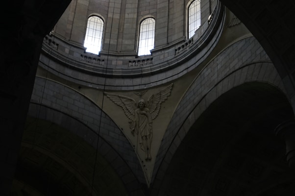 A detailed architectural interior scene with tall arched windows allowing light to enter. The intricate stone carvings include an angelic figure with wings prominently displayed against the curved surfaces of the structure.