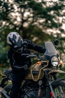 A person wearing a helmet and protective gear sits on a Royal Enfield motorcycle. The motorcycle is parked outdoors with a blurred background of trees. The scene is focused on the rider and the detailing of the motorcycle, including its rugged design and frontal headlight.