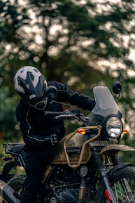 A person wearing a helmet and protective gear sits on a Royal Enfield motorcycle. The motorcycle is parked outdoors with a blurred background of trees. The scene is focused on the rider and the detailing of the motorcycle, including its rugged design and frontal headlight.
