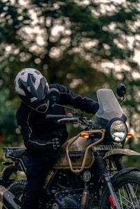 A person wearing a helmet and protective gear sits on a Royal Enfield motorcycle. The motorcycle is parked outdoors with a blurred background of trees. The scene is focused on the rider and the detailing of the motorcycle, including its rugged design and frontal headlight.