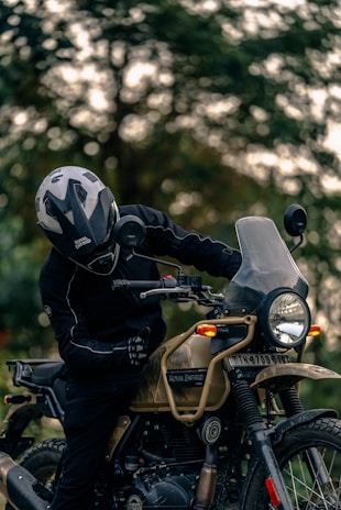 A person wearing a helmet and protective gear sits on a Royal Enfield motorcycle. The motorcycle is parked outdoors with a blurred background of trees. The scene is focused on the rider and the detailing of the motorcycle, including its rugged design and frontal headlight.