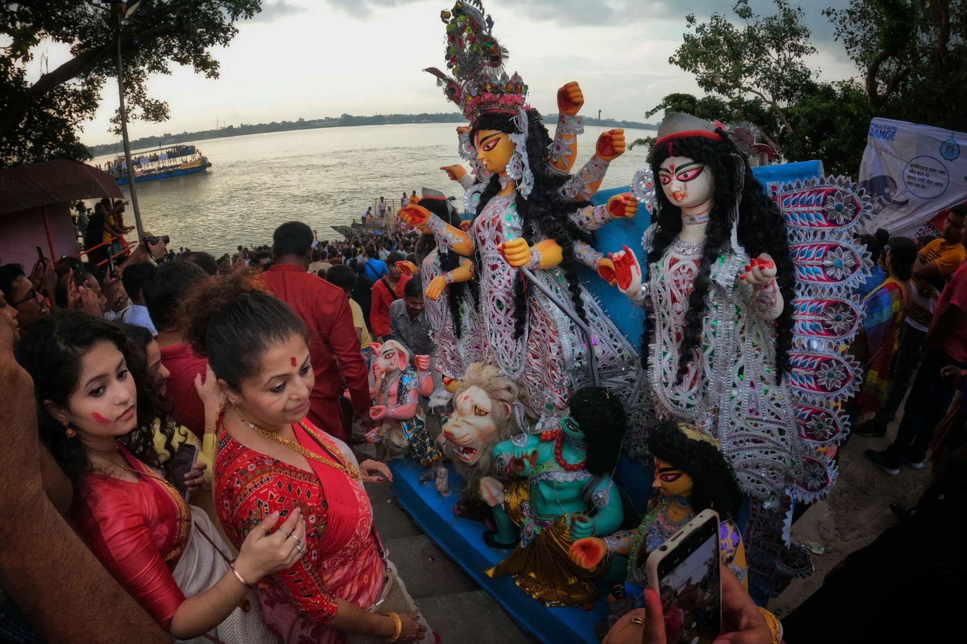 A large crowd gathers by the riverside, participating in a colorful cultural festival. People are dressed in traditional attire, and prominently displayed are intricately designed statues with vibrant colors and elaborate embellishments of a goddess figure. The atmosphere appears festive and spiritual, with many attendees actively engaging and perhaps performing religious rituals.