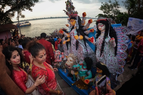 A large crowd gathers by the riverside, participating in a colorful cultural festival. People are dressed in traditional attire, and prominently displayed are intricately designed statues with vibrant colors and elaborate embellishments of a goddess figure. The atmosphere appears festive and spiritual, with many attendees actively engaging and perhaps performing religious rituals.
