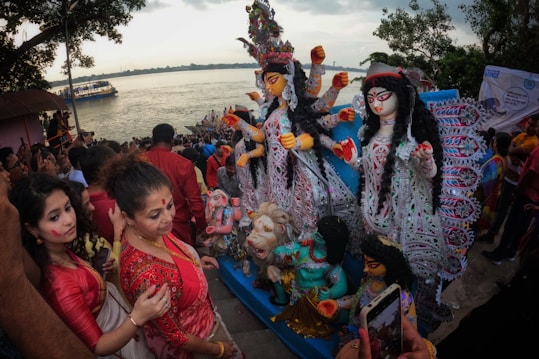 A large crowd gathers by the riverside, participating in a colorful cultural festival. People are dressed in traditional attire, and prominently displayed are intricately designed statues with vibrant colors and elaborate embellishments of a goddess figure. The atmosphere appears festive and spiritual, with many attendees actively engaging and perhaps performing religious rituals.