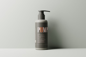 A minimalist, single pump bottle stands against a muted green background. The label is dark with bold letters spelling 'PUMP,' and fine print detailing the product.