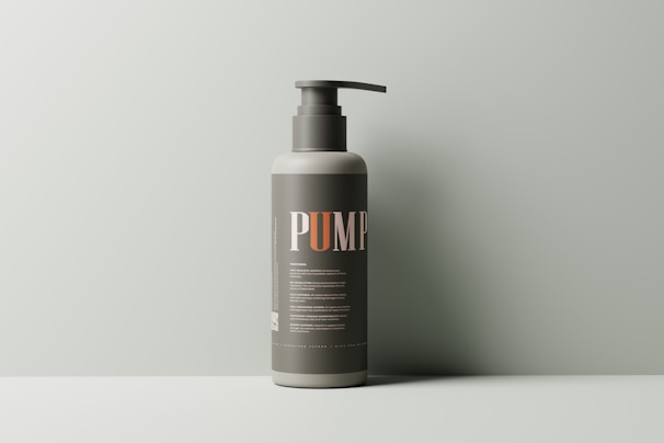 A minimalist, single pump bottle stands against a muted green background. The label is dark with bold letters spelling 'PUMP,' and fine print detailing the product.