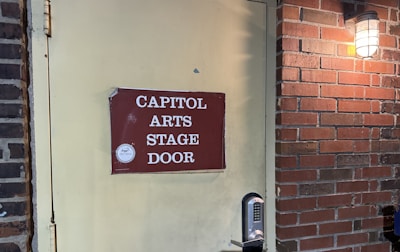 A weathered red sign with white lettering reading 'Capitol Arts Stage Door' is affixed to a pale yellow door. The door features a modern keypad lock. To the right, a brick wall is partially visible, illuminated by a wall-mounted light fixture with a frosted glass cover.
