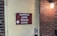 A weathered red sign with white lettering reading 'Capitol Arts Stage Door' is affixed to a pale yellow door. The door features a modern keypad lock. To the right, a brick wall is partially visible, illuminated by a wall-mounted light fixture with a frosted glass cover.