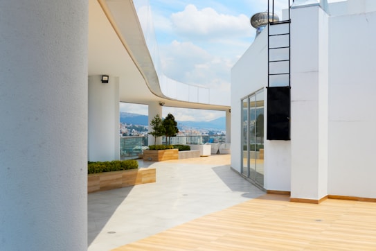 A modern rooftop terrace with clean architectural lines and a mixture of white and wooden elements. The terrace has some potted plants and a railing with glass panels that provide a view of a cityscape and mountains in the background under a partly cloudy sky. There is a black metal ladder affixed to a white structure, and the area is well-lit with natural sunlight.