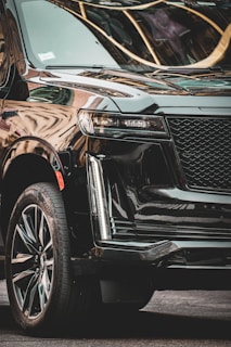 A luxurious black SUV with a shiny, sleek exterior is parked in an urban setting. The vehicle has polished alloy wheels and modern LED headlights. Reflections of nearby buildings are visible on its glossy surface.