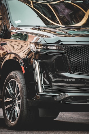 A luxurious black SUV with a shiny, sleek exterior is parked in an urban setting. The vehicle has polished alloy wheels and modern LED headlights. Reflections of nearby buildings are visible on its glossy surface.