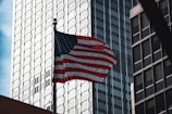 An image of the American flag gently waving against a clear sky, framed by traditional brick buildings.