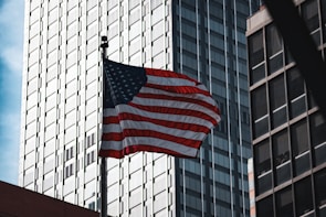 An image of the American flag gently waving against a clear sky, framed by traditional brick buildings.