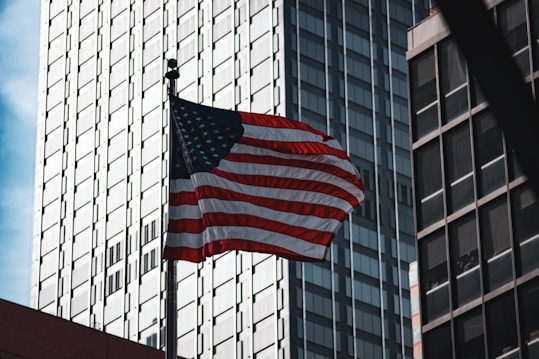 The image captures an American flag waving prominently against the backdrop of tall, modern skyscrapers with numerous windows. The flag is illuminated by sunlight, highlighting its red, white, and blue colors.